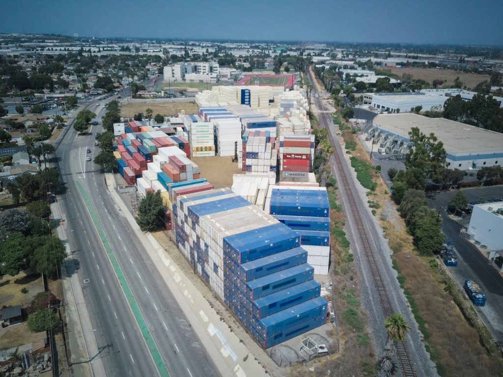 Drone aerial view of stacked cargo containers by roads and train tracks in an urban area.
